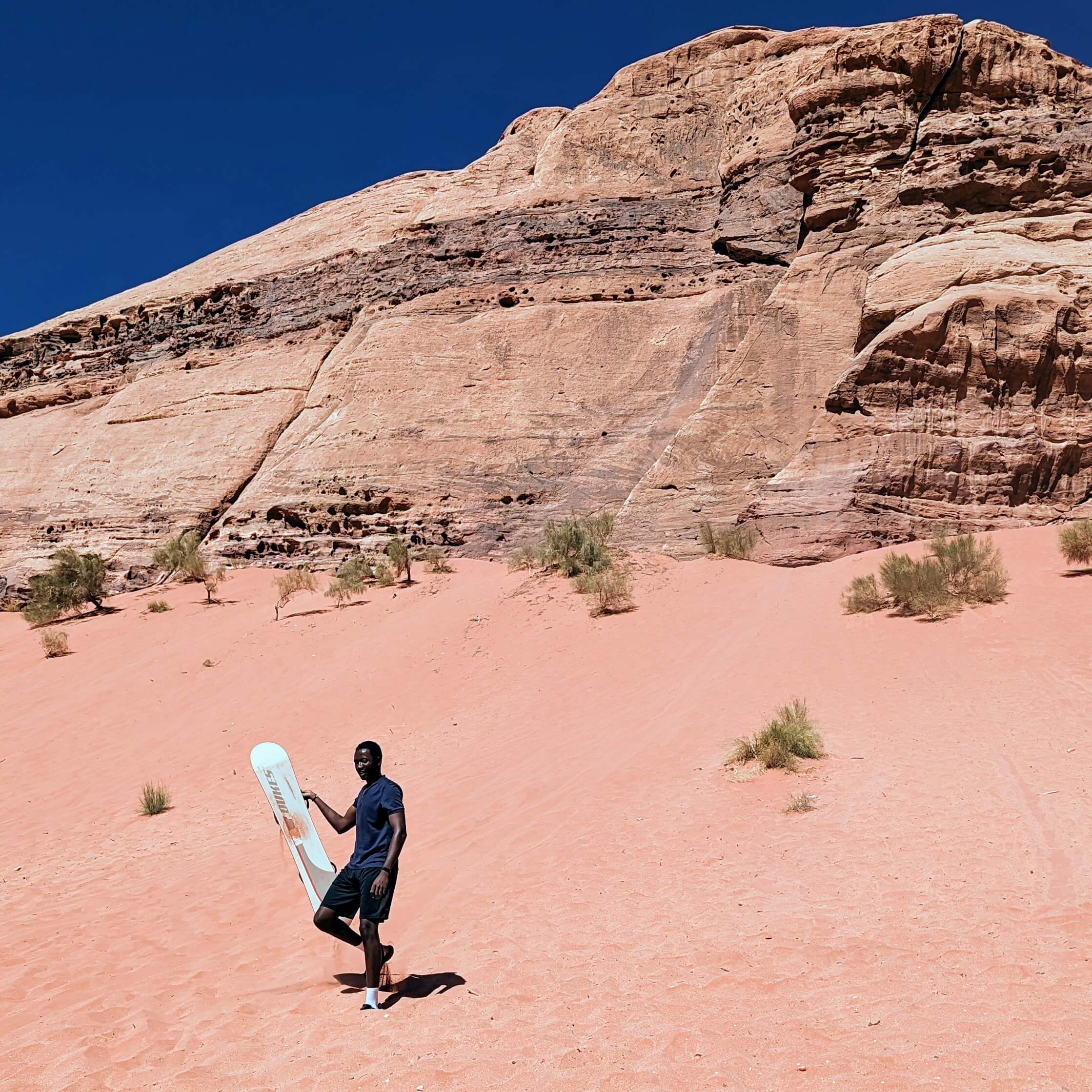 Holding a sandboard in a sandy area with mountains looming in the background